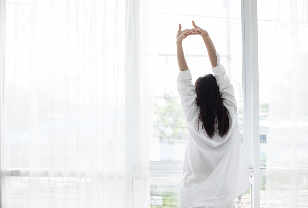Mulher asiática acordando em sua cama totalmente descansada e abre as cortinas de manhã para tomar ar fresco.