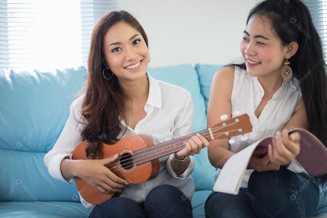 Duas mulheres asiáticas estão se divertindo tocando ukulele e sorrindo em casa para relaxar