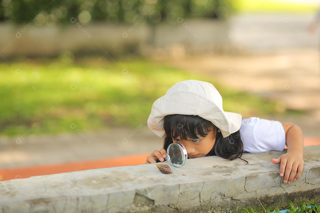 Menina asiática vestindo um chapéu branco e explorando a natureza com uma lupa.