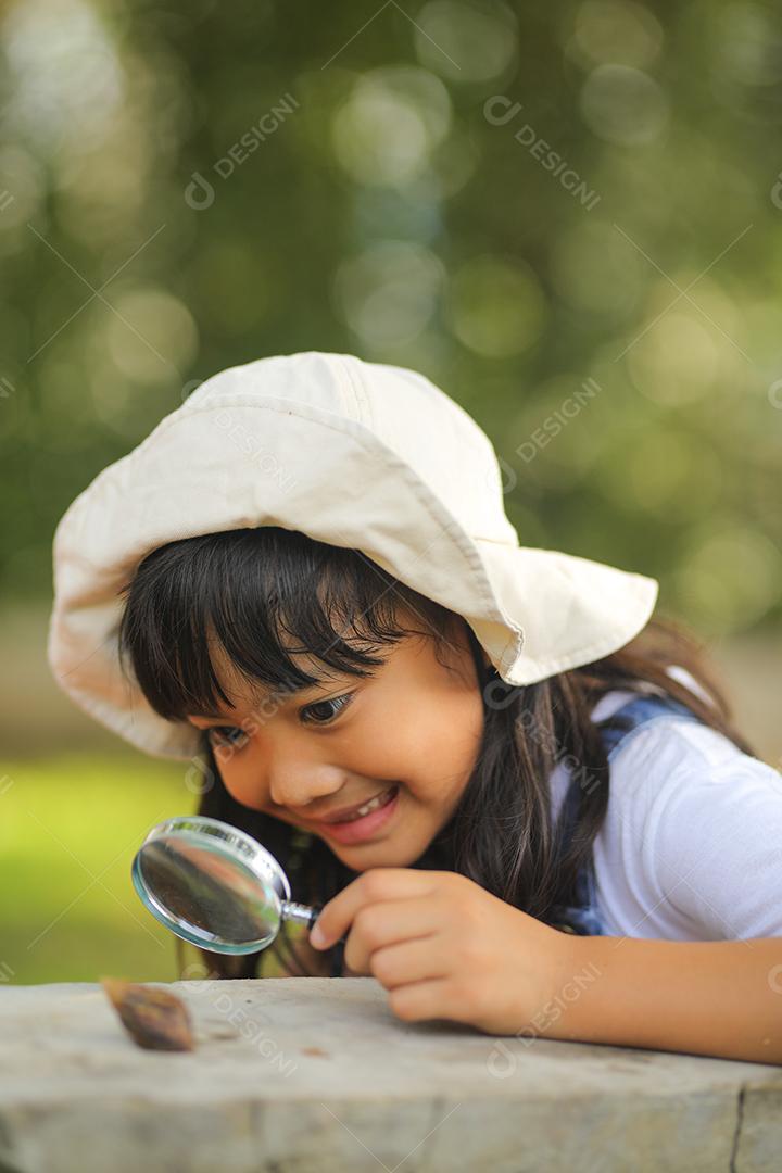 Menina asiática vestindo um chapéu branco e explorando a natureza com uma lupa.