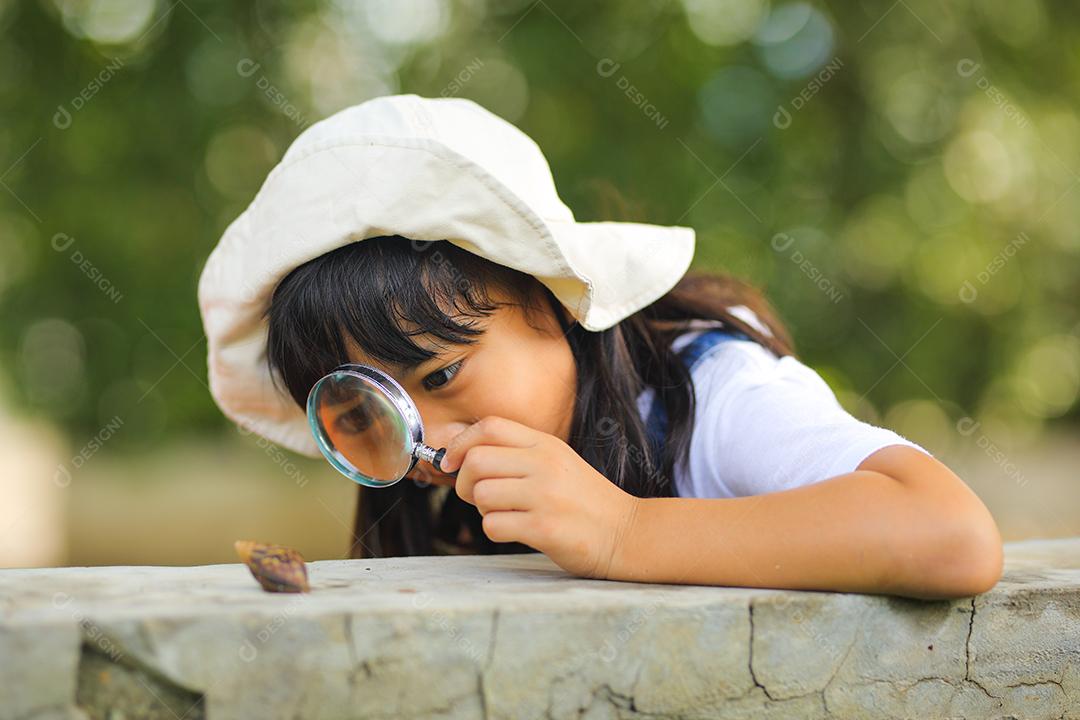 Menina asiática vestindo um chapéu branco e explorando a natureza com uma lupa.