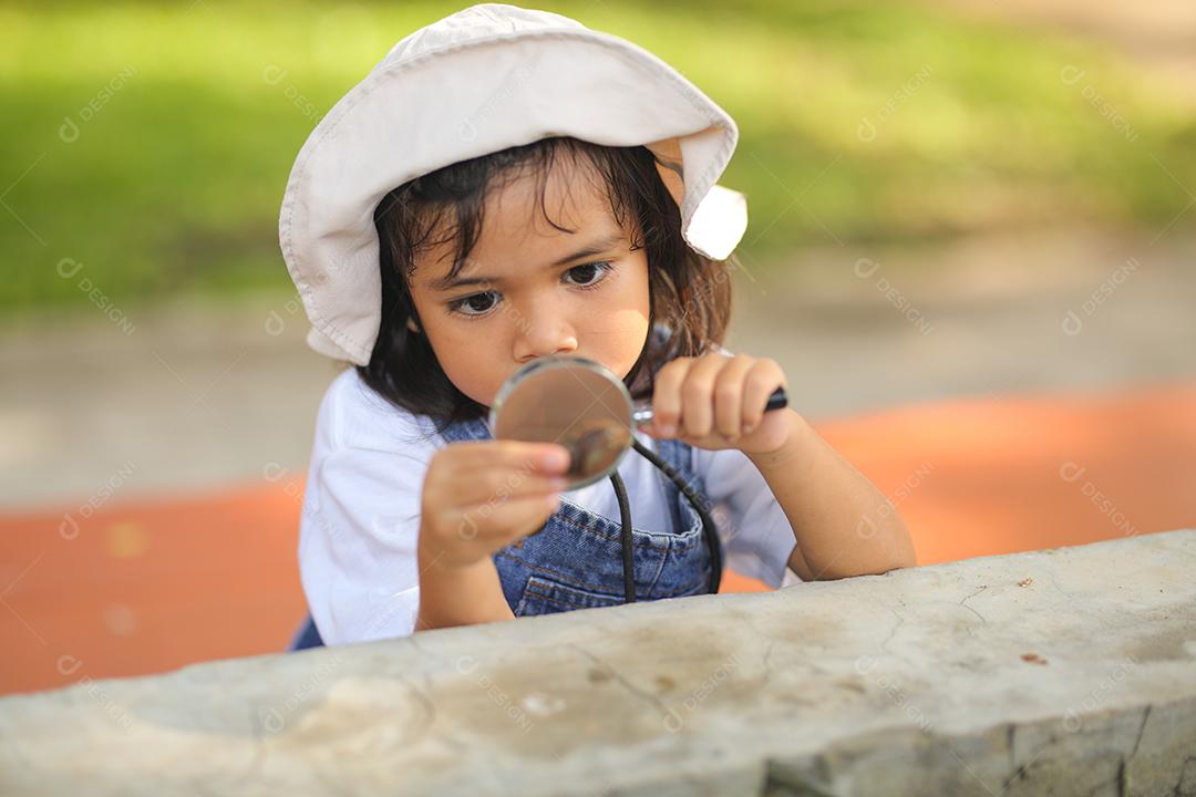 Menina asiática vestindo um chapéu branco e explorando a natureza com uma lupa.