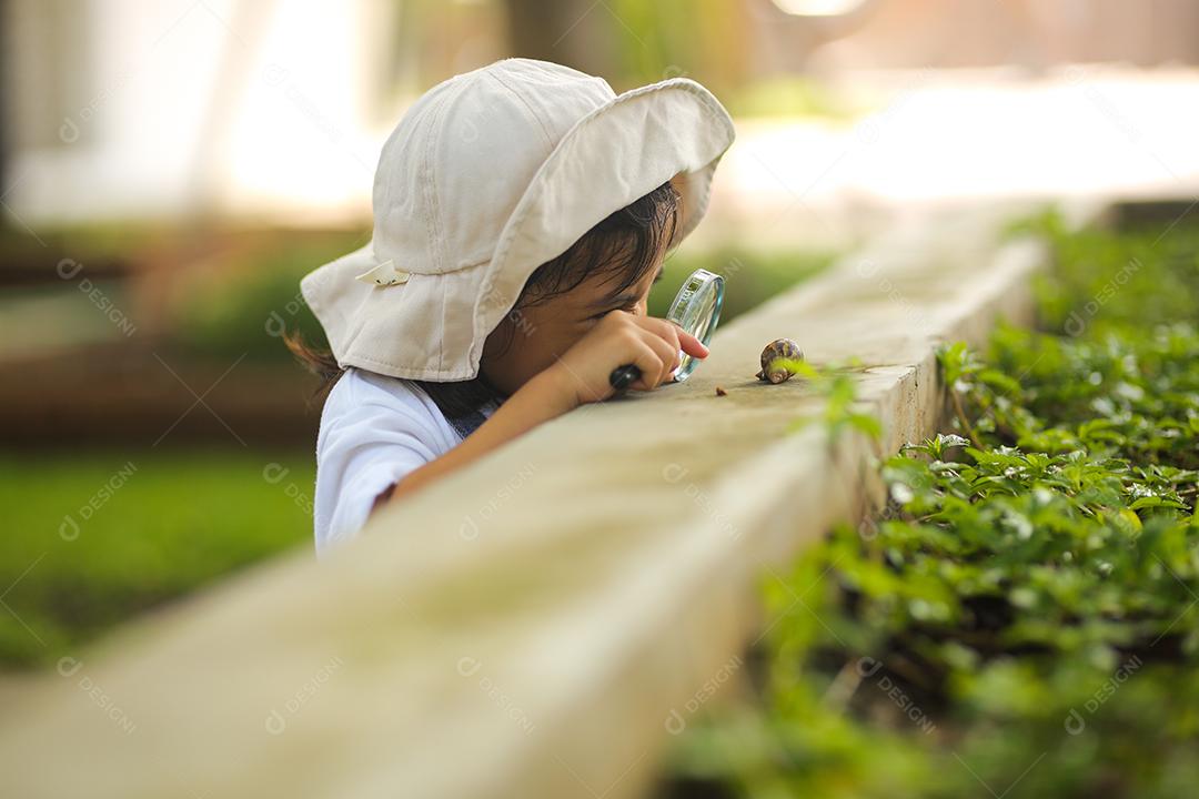 Menina asiática vestindo um chapéu branco e explorando a natureza com uma lupa.