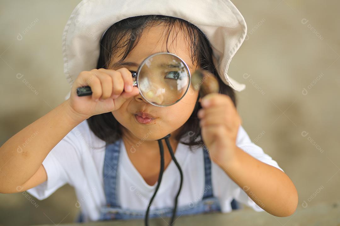 Menina asiática vestindo um chapéu branco e explorando a natureza com uma lupa.