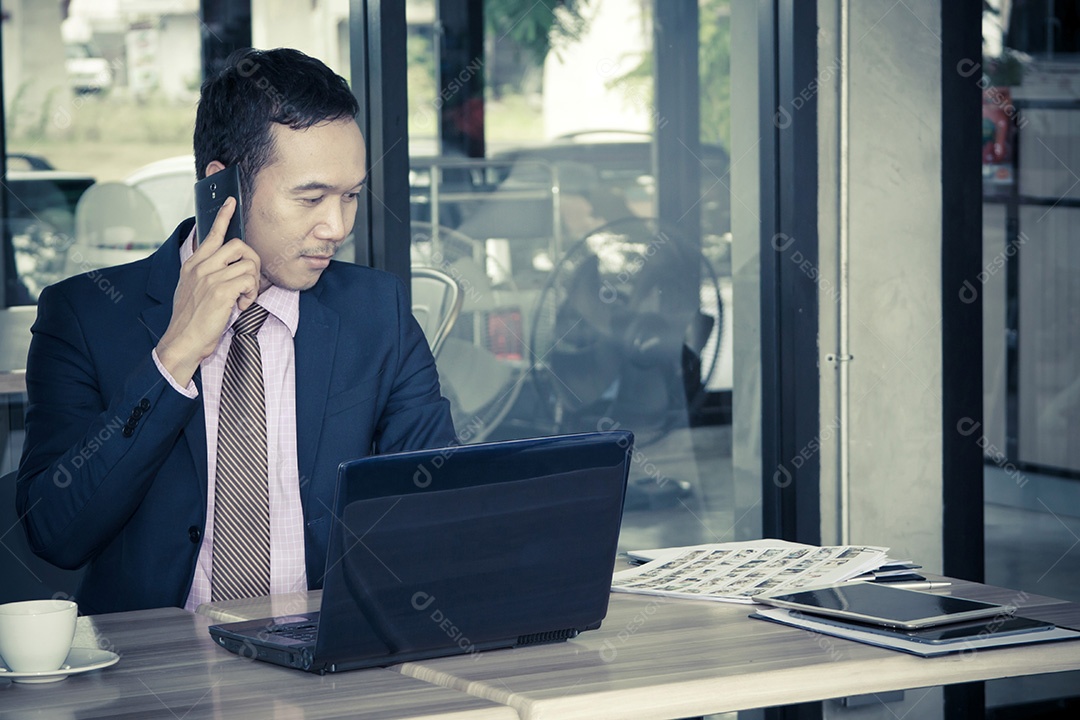 Jovem empresário sentado no café, falando no celular, com laptop