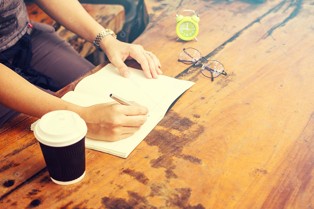 Mãos com caneta escrevendo no caderno, café e relógio