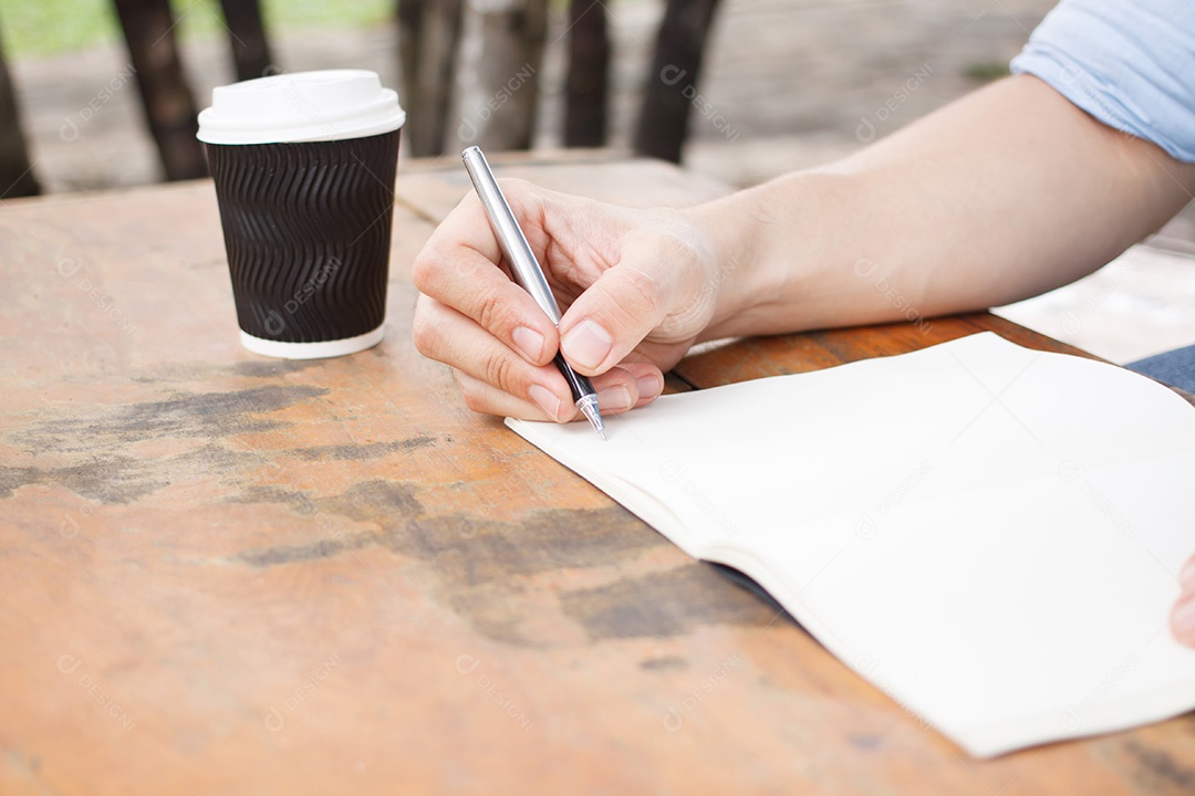 Mãos com caneta escrevendo no caderno com café no parque