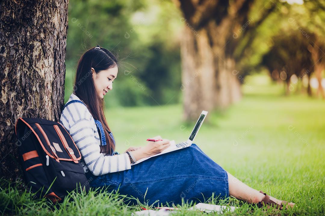 Linda garota asiática sorridente lendo livro e trabalhando na árvore no parque no verão para relaxar