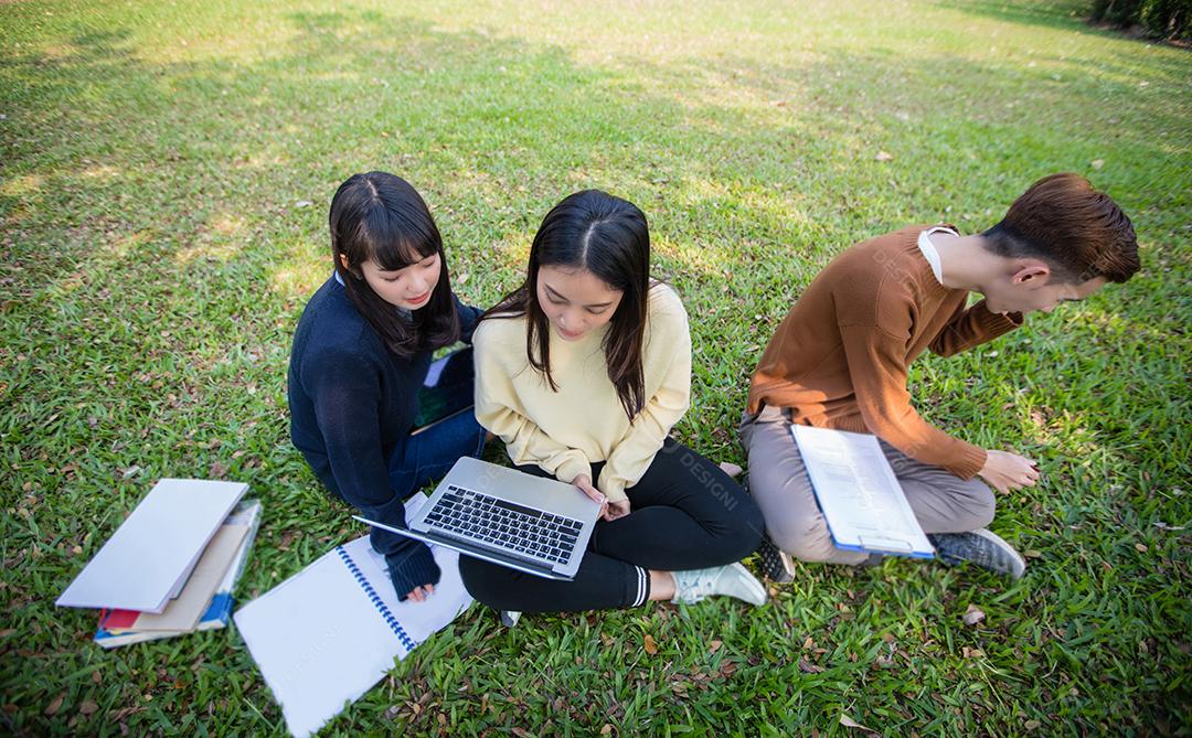 Grupo de estudantes universitários asiáticos sentados na grama verde trabalhando e lendo fora juntos em um parque