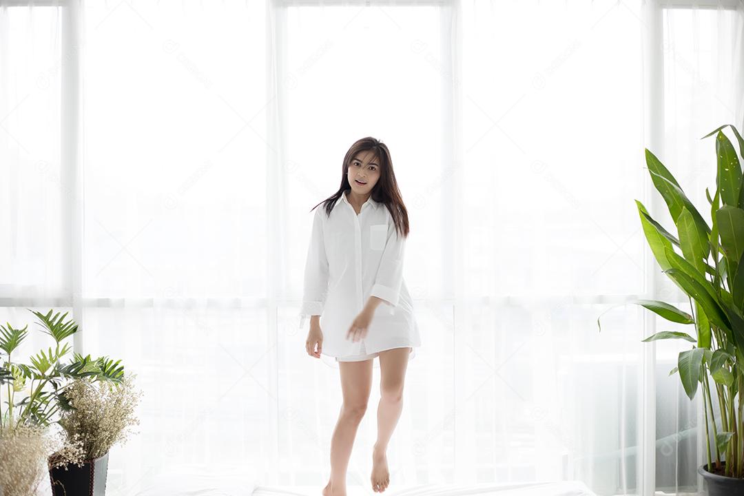 Mulher asiática acorda em sua cama totalmente descansada e abre as cortinas de manhã para tomar ar fresco.