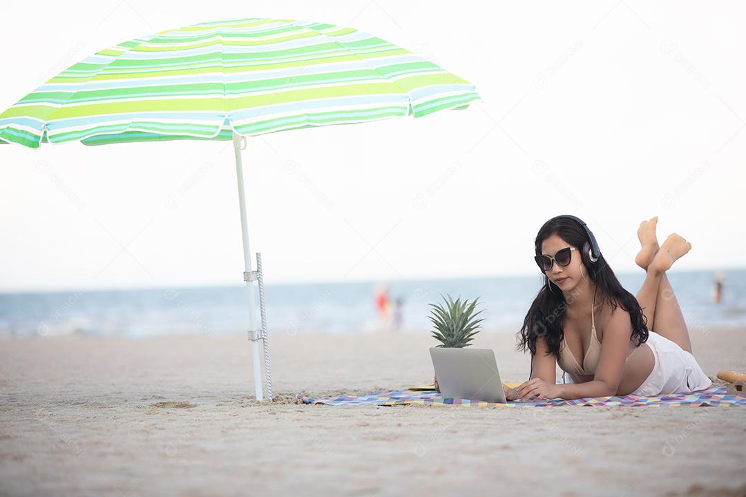 Mulher vestindo biquínis estão usando laptop para trabalhar e ouvir música na praia no verão.