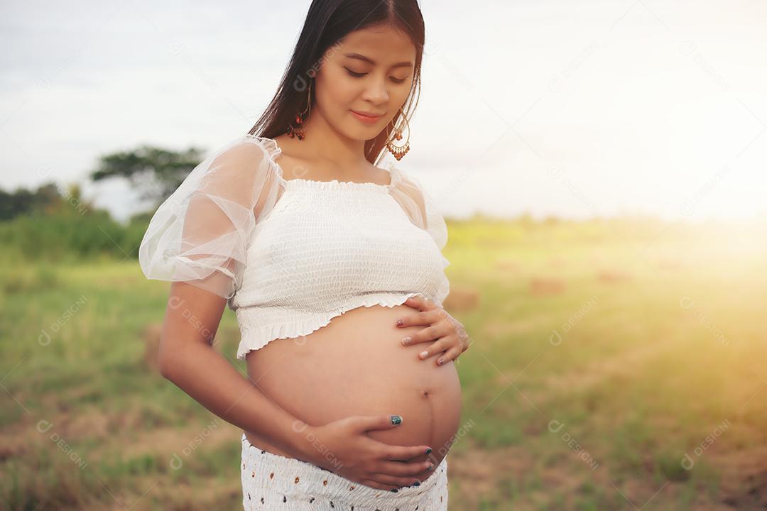 happy and proud asian pregnant woman looking at her belly in a park at sunrise