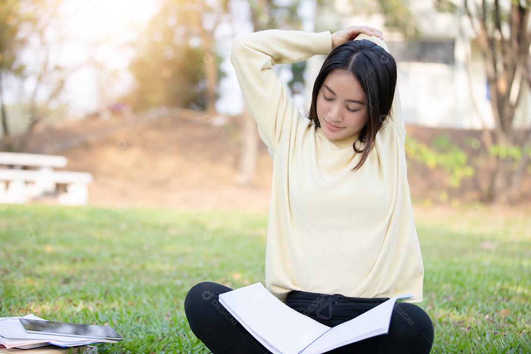 Mulher asiática esticando e sentada na grama verde depois de ler o livro e trabalhar duro em um parque