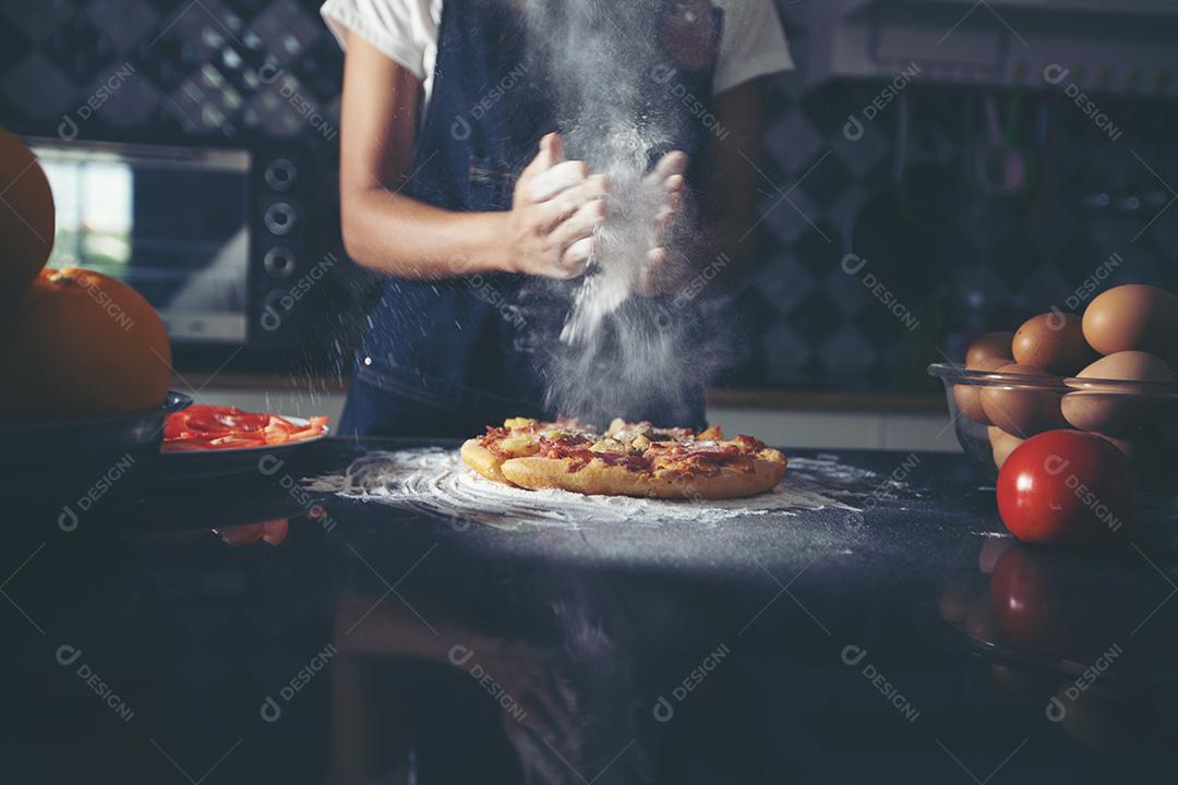 Mulheres asiáticas preparando uma pizza, amassando a massa e colocando ingredientes na mesa da cozinha