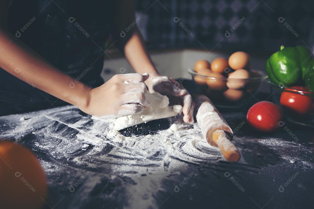 Mulheres asiáticas preparando uma pizza, amassando a massa e colocando ingredientes na mesa da cozinha