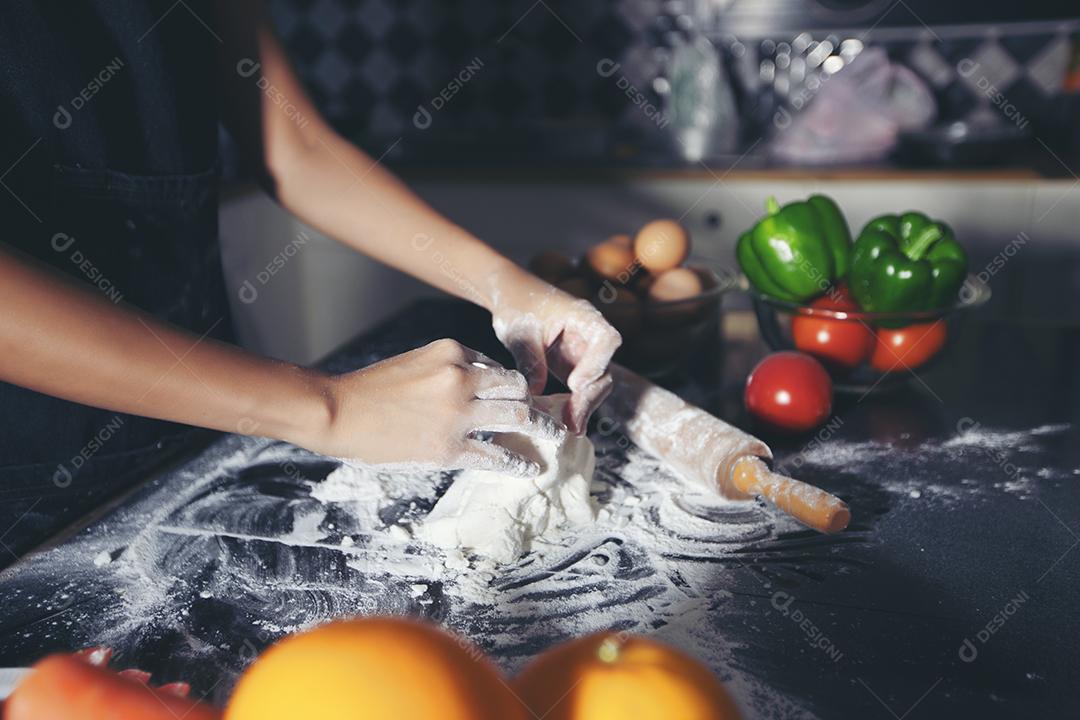 Mulheres asiáticas preparando uma pizza, amassando a massa e colocando ingredientes na mesa da cozinha