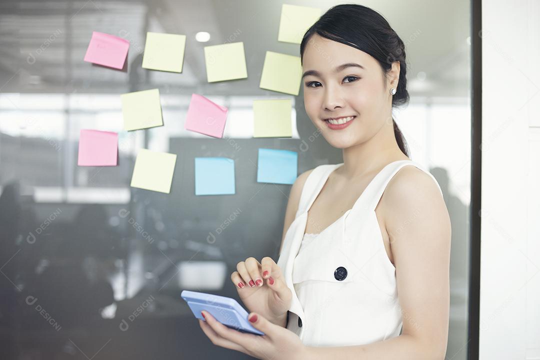 Mulheres de negócios asiáticos sorrindo e trabalhando juntos em vidro de parede com adesivos post-it. Escritório de inicialização moderno