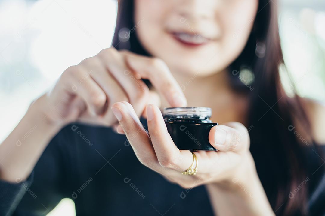 Uma linda mulher asiática usando um produto de cuidados com a pele, hidratante ou loção cuidando de sua pele seca. Creme hidratante nas mãos femininas.