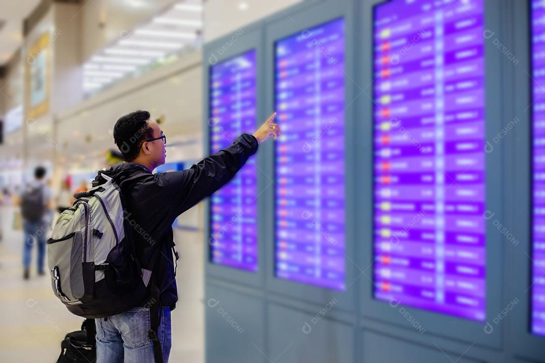 Homem asiático com viajante de mochila usando o telefone celular inteligente para check-in na tela de informações de voo em um aeroporto moderno