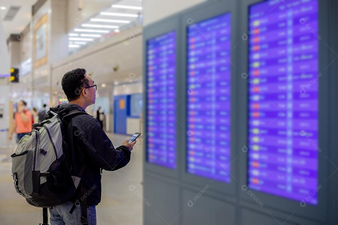 Homem asiático com viajante de mochila usando o telefone celular inteligente para check-in na tela de informações de voo em um aeroporto moderno