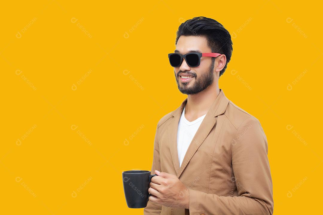 Retrato de um jovem empresário asiático bonito segurando uma xícara e bebendo café. Isolado em fundo amarelo.