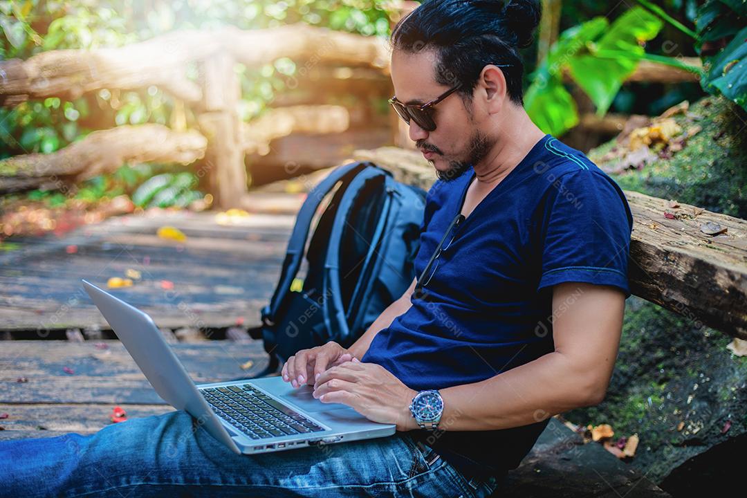 Homem asiático trabalhando e verificando fotos no laptop na natureza ao ar livre e ele está relaxando, desfrutando de viagens