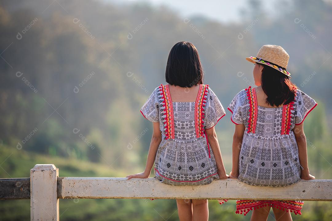 Duas garotas asiáticas bonitas e jovens sentadas na cerca do jardim no pôr do sol