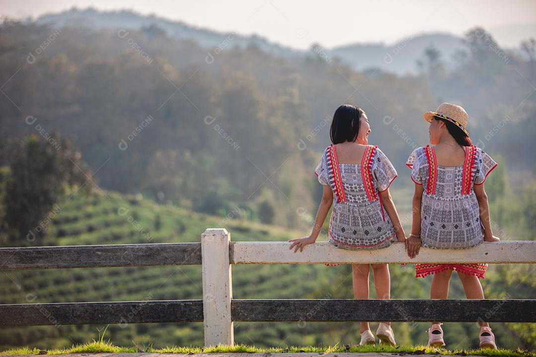 Duas garotas asiáticas bonitas e jovens sentadas na cerca do jardim no pôr do sol