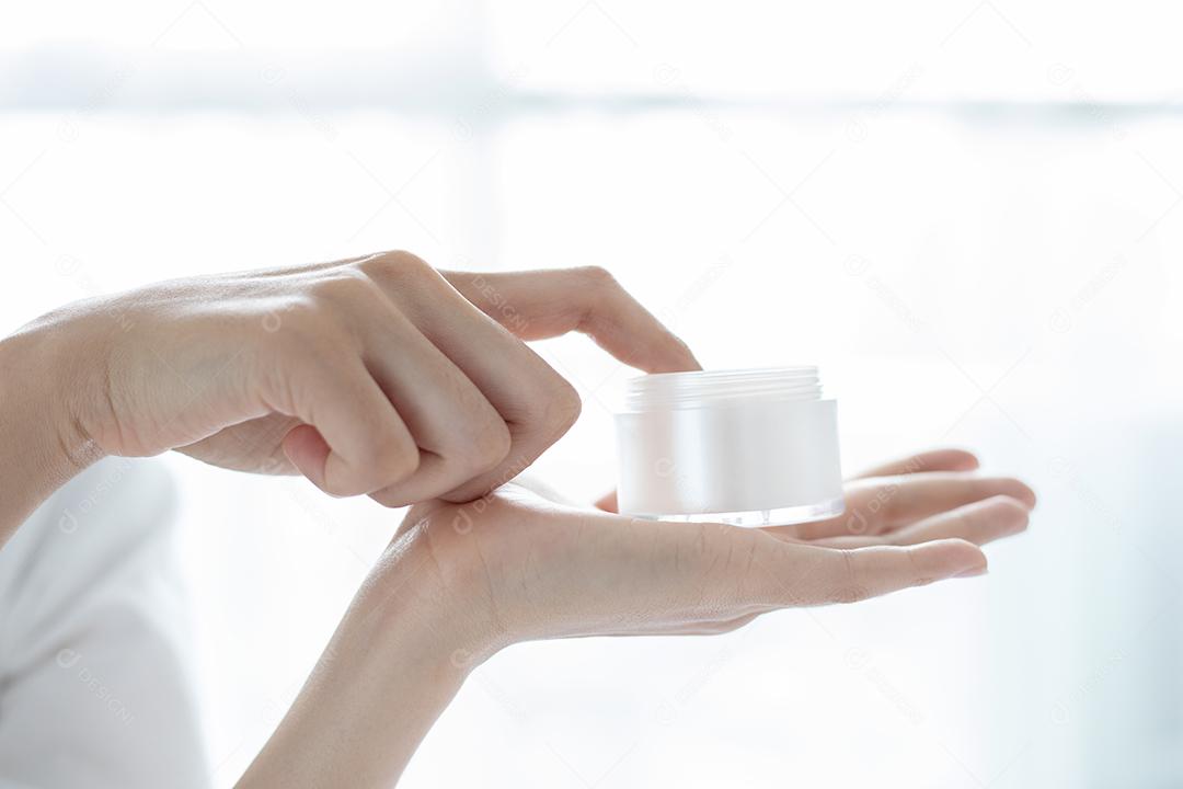 Uma linda mulher asiática usando um produto de cuidados com a pele, hidratante ou loção cuidando de sua pele seca. Creme hidratante nas mãos femininas.