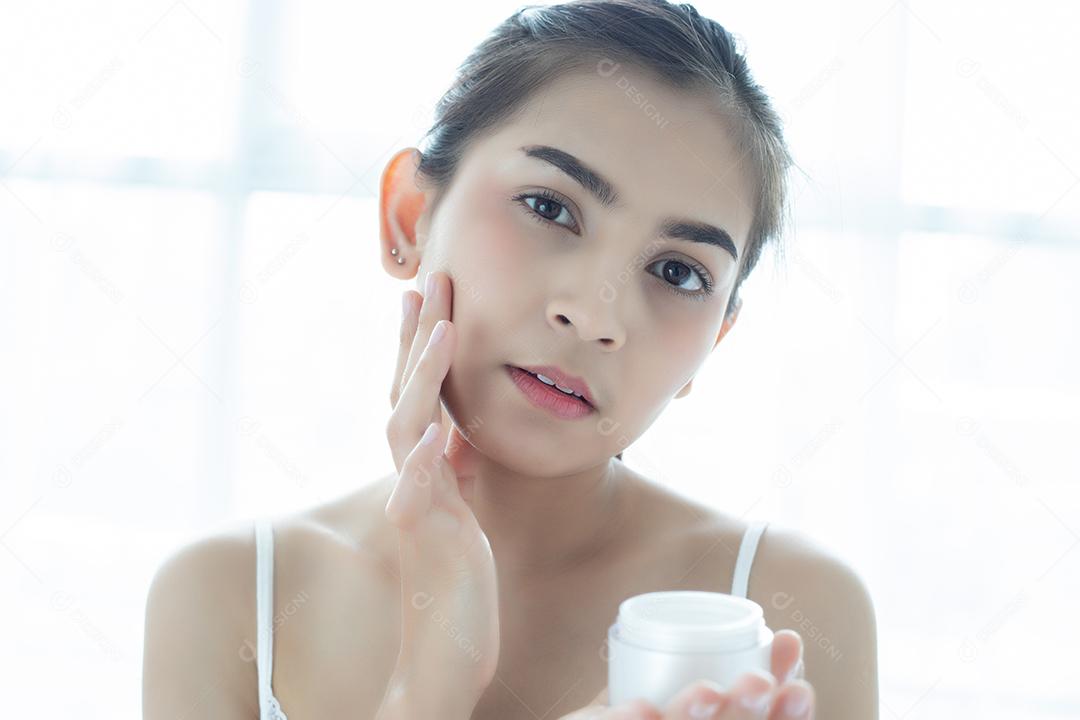 Uma linda mulher asiática usando um produto de cuidados com a pele, hidratante ou loção cuidando de sua pele seca. Creme hidratante nas mãos femininas.