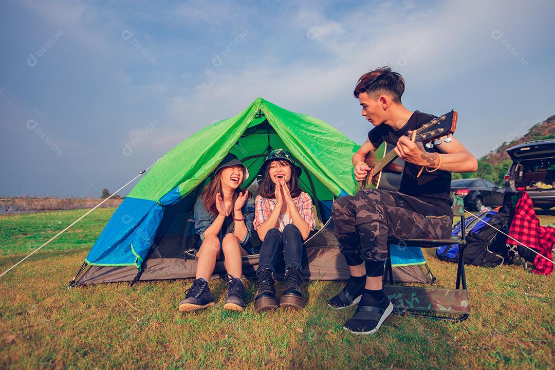 Um grupo de amigos asiáticos turista bebendo e tocando violão junto com felicidade no verão enquanto acampa perto do lago ao pôr do sol