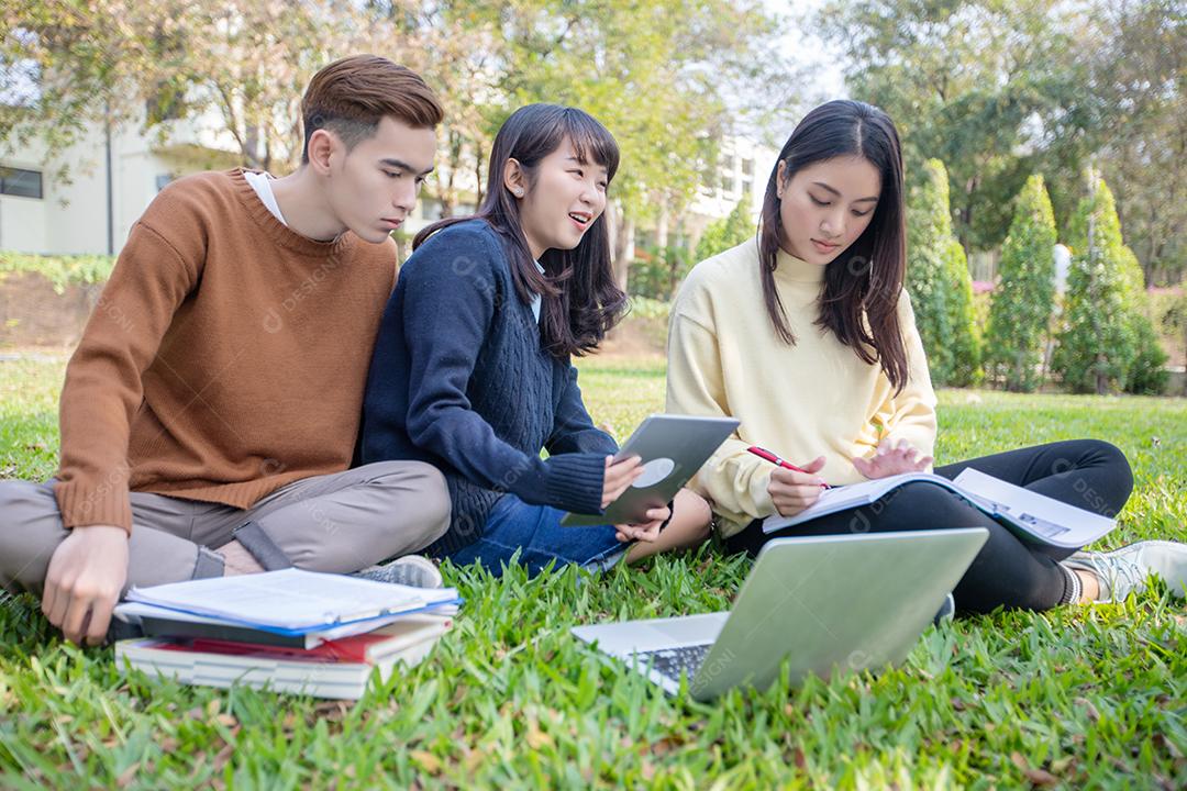 Grupo de estudantes universitários asiáticos sentados na grama verde trabalhando e lendo fora juntos em um parque