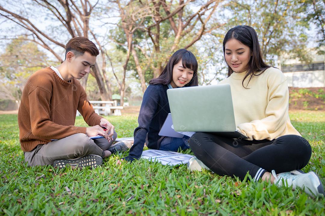 Grupo de estudantes universitários asiáticos sentados na grama verde trabalhando e lendo fora juntos em um parque