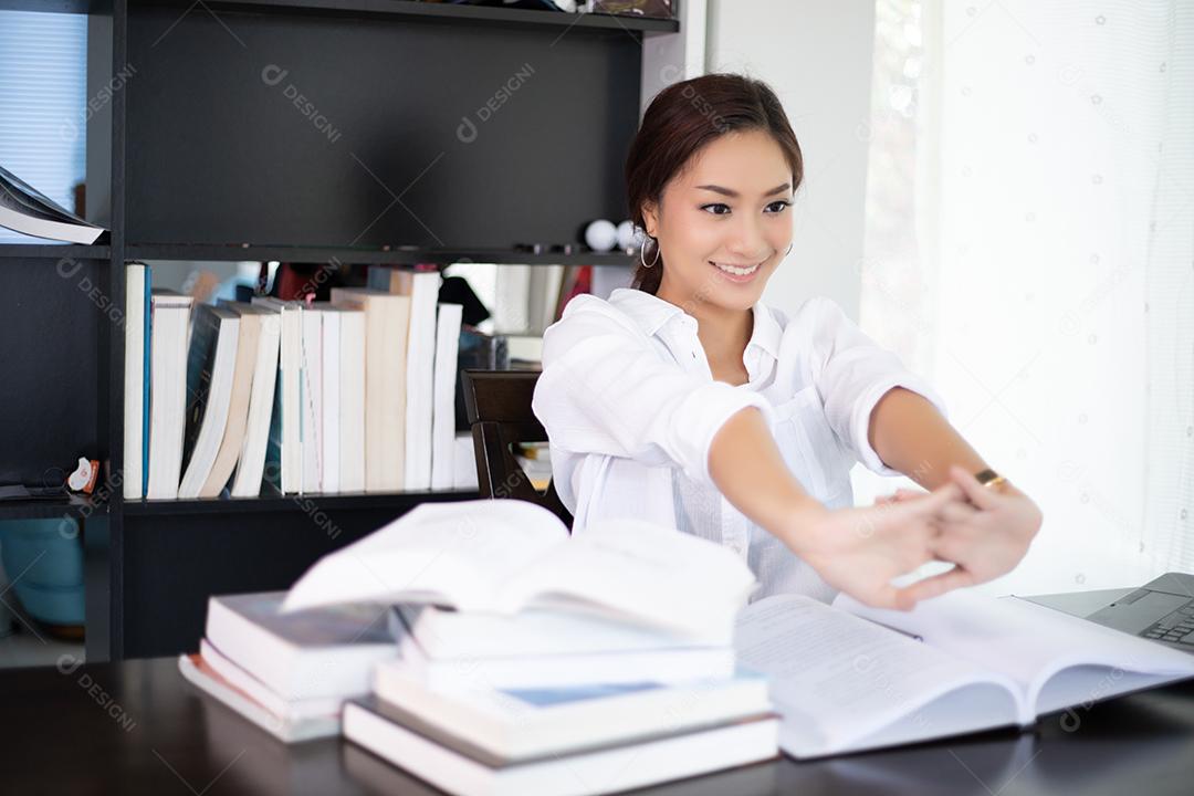 Mulher asiática esticando depois de ler o livro e trabalhar duro e sorrindo no escritório em casa