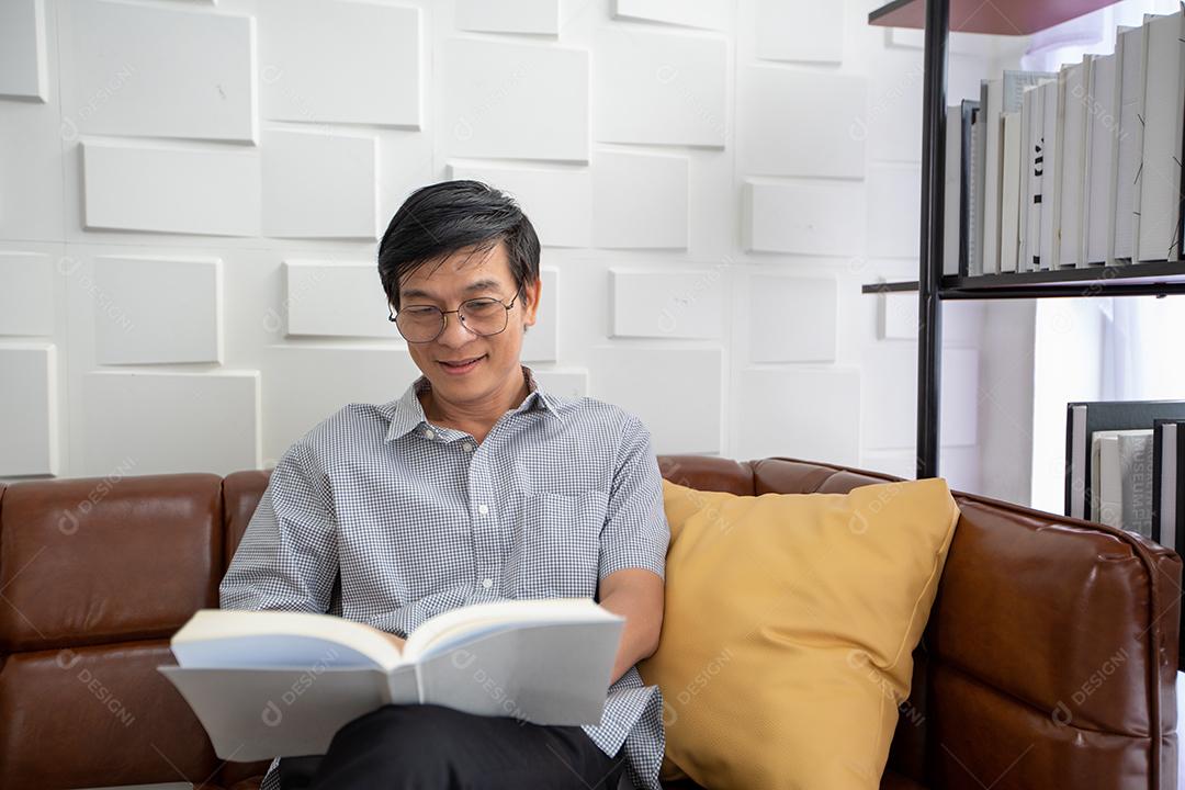 Homem asiático sênior lendo livro no sofá na sala de estar em casa, retrato de homem idoso asiático é relaxante e felicidade com ler uma revista