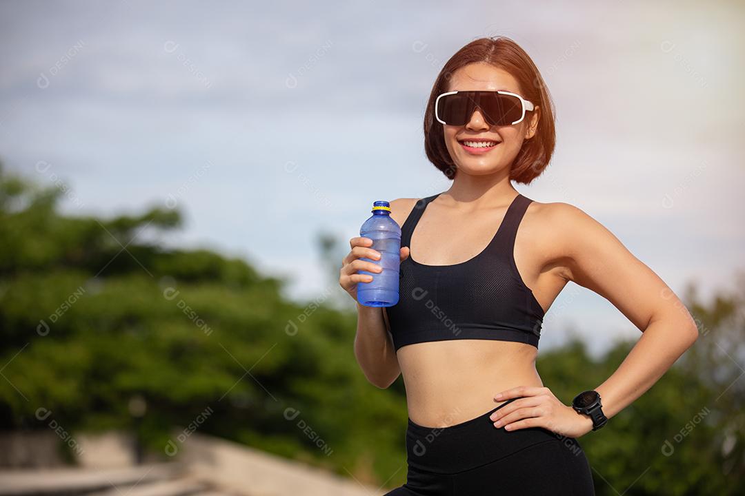 Mulher esportiva asiática bebendo água ao ar livre depois de correr em dia ensolarado