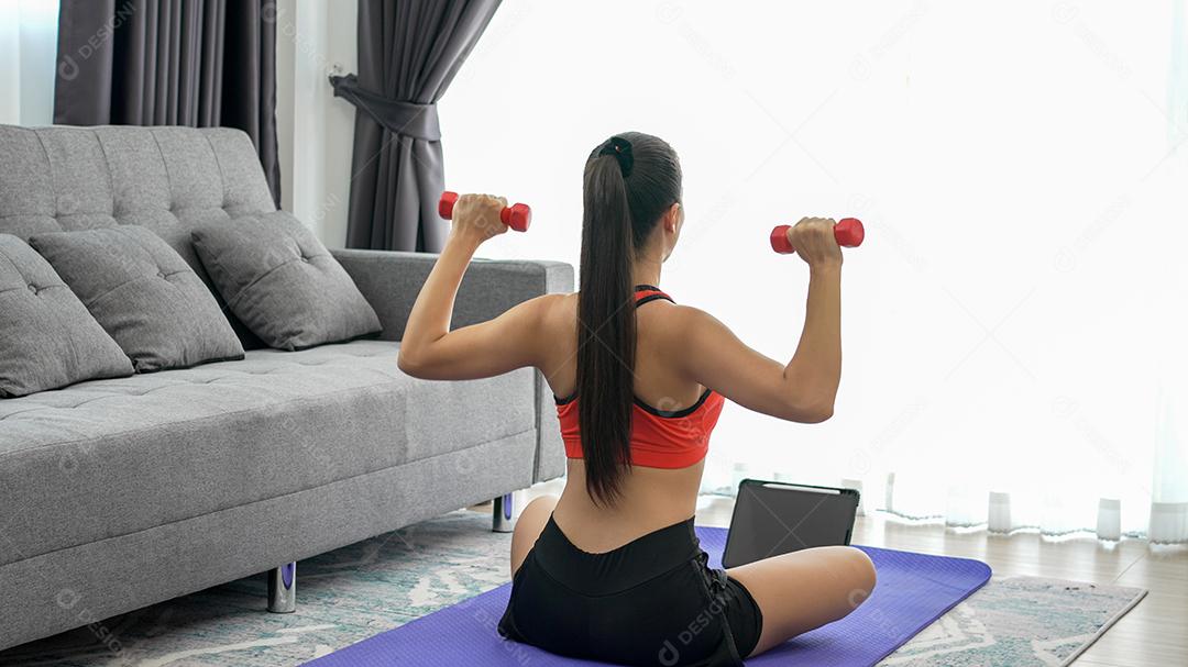 Atleta jovem fazendo exercício com haltere no tapete de ioga com tablet em casa