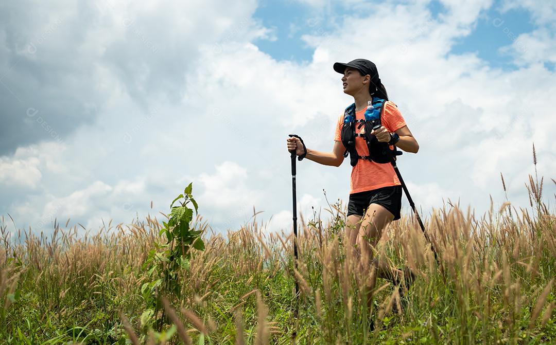 Trilha ativa de mulheres jovens atravessando um prado em uma trilha gramada no alto das montanhas à tarde com vara de trekking