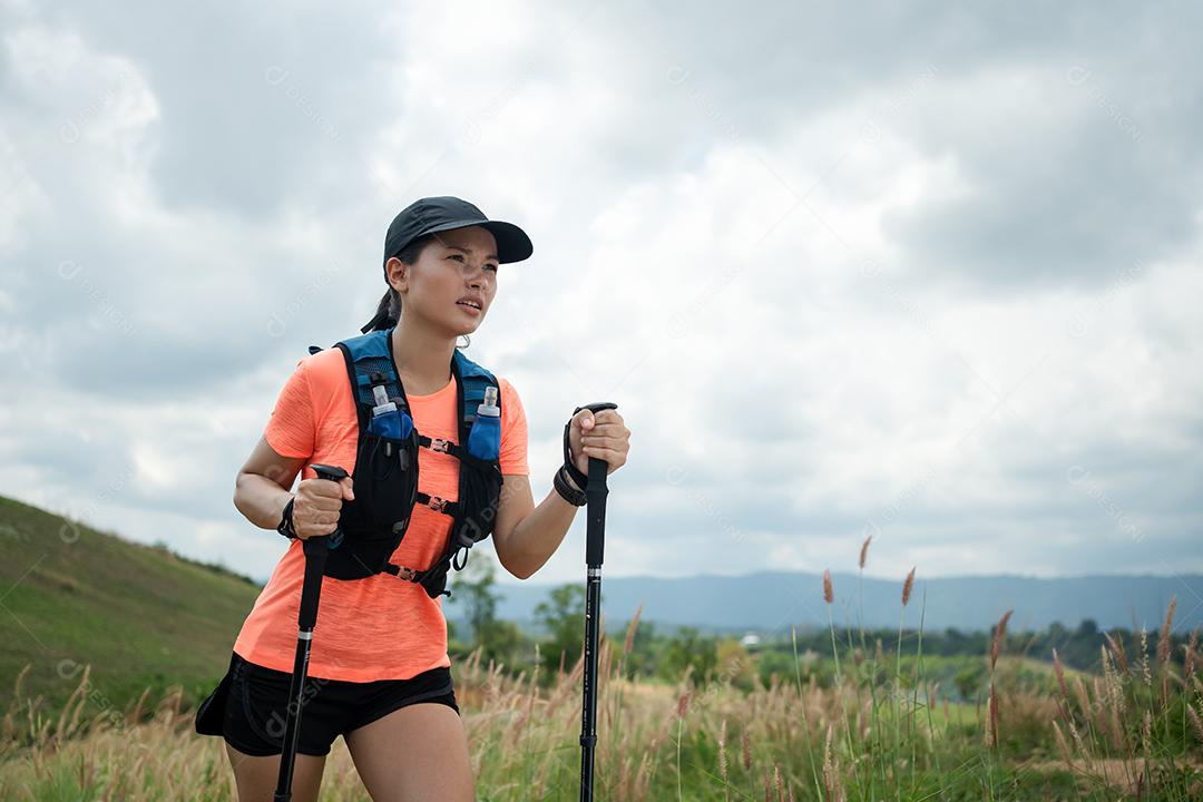 Trilha ativa de mulheres jovens atravessando um prado em uma trilha gramada no alto das montanhas à tarde com vara de trekking