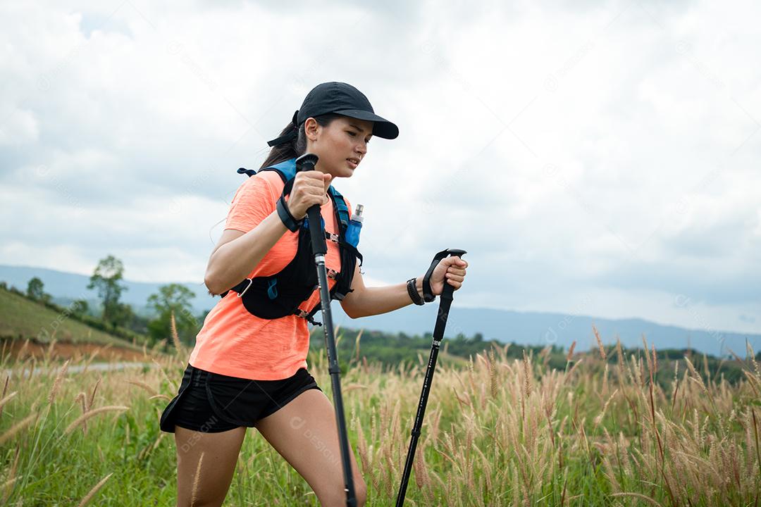 Trilha ativa de mulheres jovens atravessando um prado em uma trilha gramada no alto das montanhas à tarde com vara de trekking