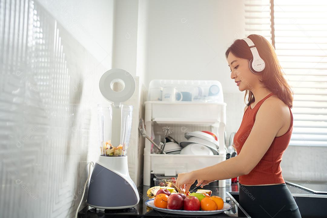 A desportista fazendo smoothies de frutas na cozinha em casa enquanto ouve música através de fones de ouvido - conceitos de estilo de vida