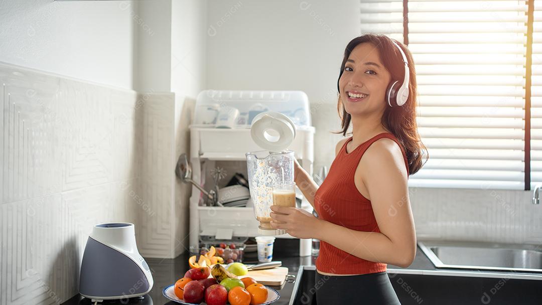 Mulher jovem e bonita fazendo e bebendo smoothies de frutas na cozinha em casa enquanto ouve música através de fones de ouvido - conceitos de estilo de vida