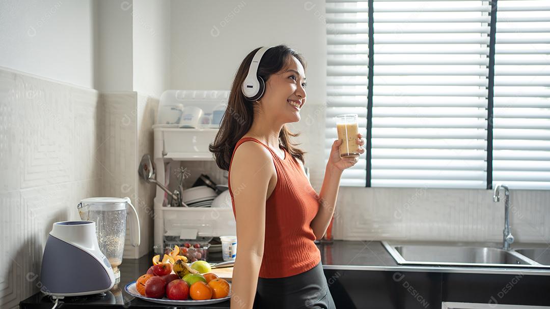 Mulher jovem e bonita fazendo e bebendo smoothies de frutas na cozinha em casa enquanto ouve música através de fones de ouvido - conceitos de estilo de vida