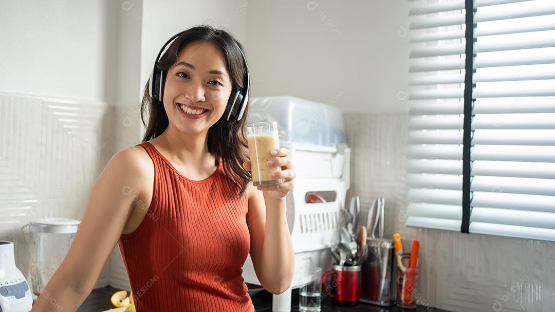 Mulher jovem e bonita fazendo e bebendo smoothies de frutas na cozinha em casa enquanto ouve música através de fones de ouvido - conceitos de estilo de vida