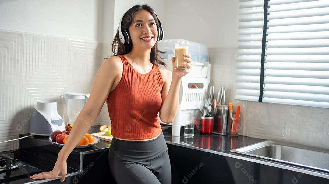 Mulher jovem e bonita fazendo e bebendo smoothies de frutas na cozinha em casa enquanto ouve música através de fones de ouvido - conceitos de estilo de vida