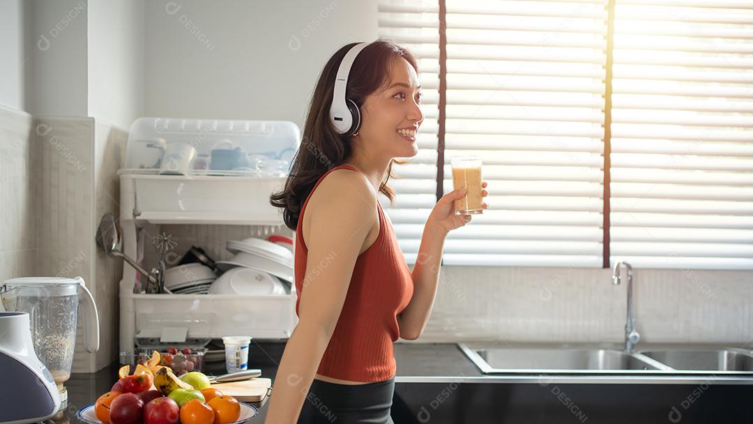 Mulher jovem e bonita fazendo e bebendo smoothies de frutas na cozinha em casa enquanto ouve música através de fones de ouvido - conceitos de estilo de vida