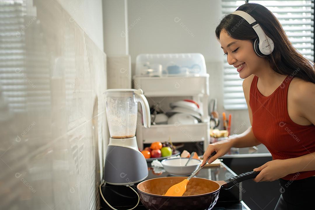 Linda jovem cozinhando na cozinha em casa enquanto ouve música através de fones de ouvido - conceitos de estilo de vida