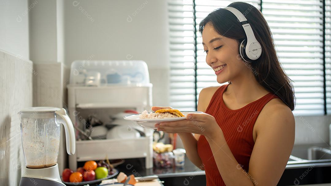 Linda jovem cozinhando na cozinha em casa enquanto ouve música através de fones de ouvido - conceitos de estilo de vida