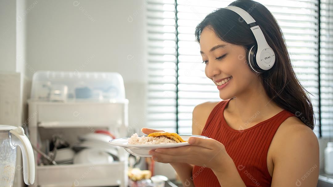 Linda jovem cozinhando na cozinha em casa enquanto ouve música através de fones de ouvido - conceitos de estilo de vida