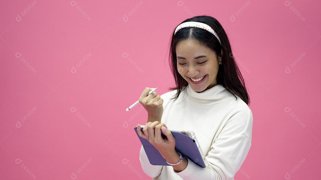 Mulher asiática sorrindo e usando o celular com tablet e animada para fazer compras online em fundo rosa isolado.